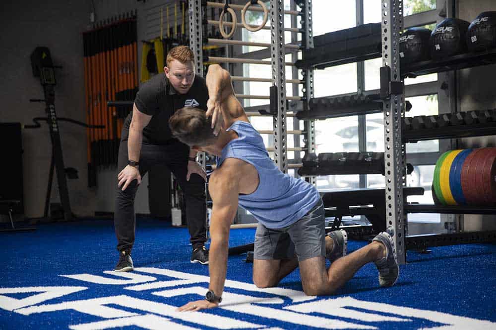 mobility expert adjusts shoulder of client kneeling in gym performing a half twist with one hand on the ground