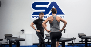 assisted stretching professional demonstrates exercise technique to client. Both pose balanced on one foot hands at hips